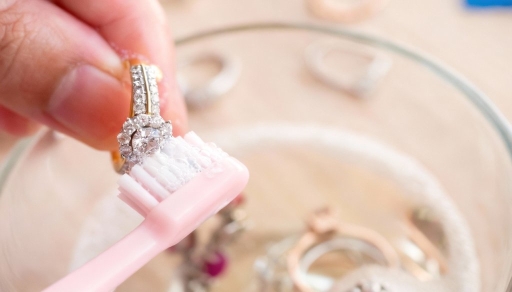 Közelről egy kéz egy gyémántgyűrűt tisztít fehér kefével, háttérben egy átlátszó edény folyadékkal látható.