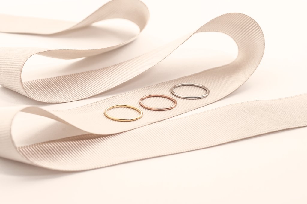 Három vékony gyűrű (sárga arany, rozé arany és fehér arany) egy világos bézs szalagon elrendezve, letisztult, elegáns stílusú, világos háttér előtt.